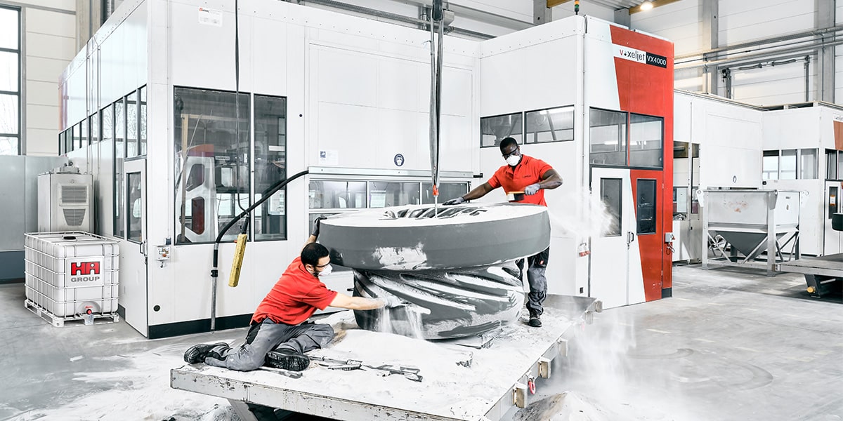 Two voxeljet employees unpack a large impeller casting core in front of a voxeljet vx4000 3d printer. Next to the printer stands a IBC container with the logo of Huettens Albertus.